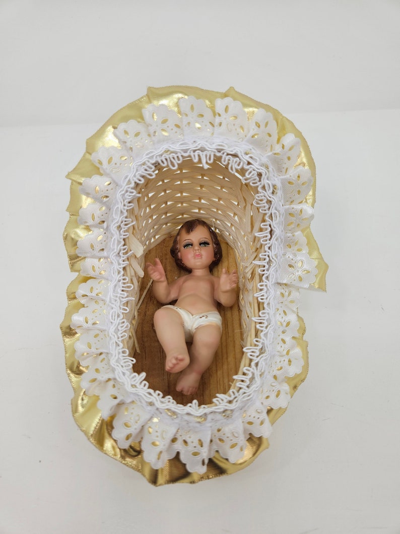 K&ouml;nnte beinhalten: Eine kleine Babyfigur mit braunen Haaren und gr&uuml;nen Augen, bekleidet mit einer wei&szlig;en Windel, liegt in einer geflochtenen Korbwiege. Die Wiege ist mit einer ger&uuml;schten wei&szlig;en Spitzenborte und einem gl&auml;nzenden goldenen Stoffrand verziert.