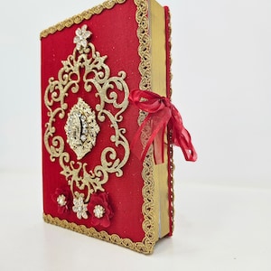 May include: A red book with gold-colored edging and a decorative gold filigree design on the cover. The cover also features a jeweled embellishment and red floral accents. A red ribbon is tied around the book.