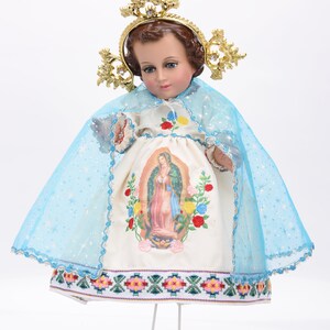 Puede incluir: Una mu&ntilde;eca de porcelana blanca vestida con un vestido blanco con velo azul y corona dorada. El vestido tiene una imagen bordada de la Virgen Mar&iacute;a y un estampado floral colorido. La mu&ntilde;eca est&aacute; de pie sobre un soporte blanco.