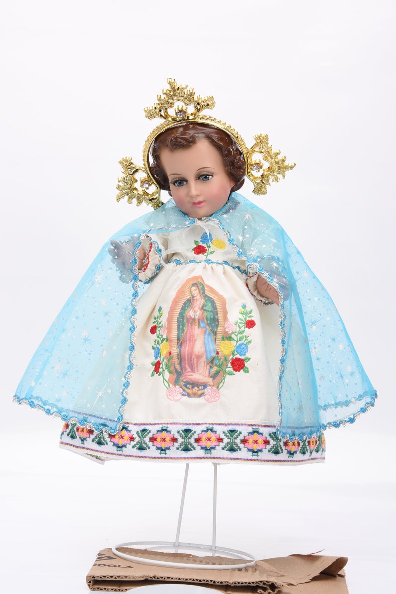 Puede incluir: Una mu&ntilde;eca de porcelana blanca vestida con un vestido blanco con velo azul y corona dorada. El vestido tiene una imagen bordada de la Virgen Mar&iacute;a y un estampado floral colorido. La mu&ntilde;eca est&aacute; de pie sobre un soporte blanco.
