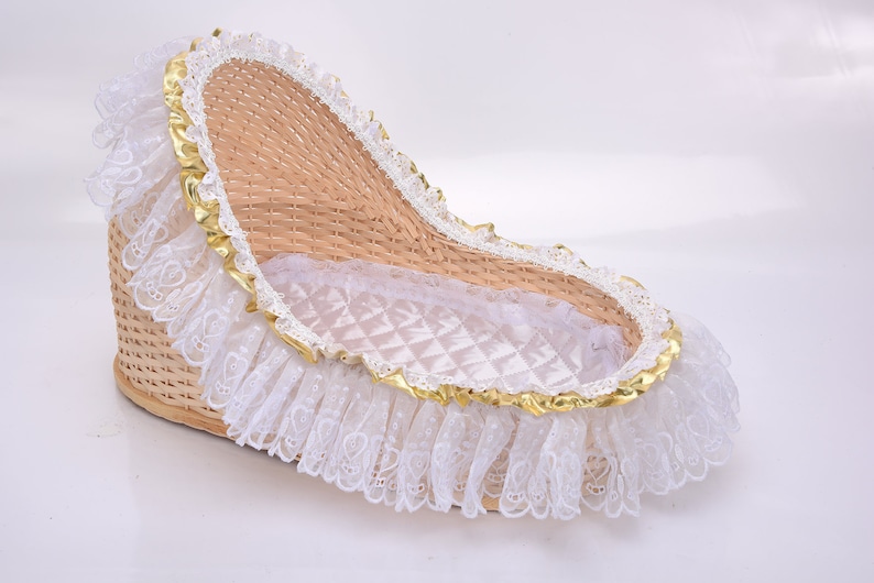 K&ouml;nnte beinhalten: Ein geflochtener Weidenkorb mit hoher R&uuml;ckenlehne und niedriger Vorderseite, ausgekleidet mit wei&szlig;em, gestepptem Stoff. Der Rand ist mit einer goldenen Metallborte und einer breiten, ger&uuml;schten wei&szlig;en Spitzenbord&uuml;re verziert.