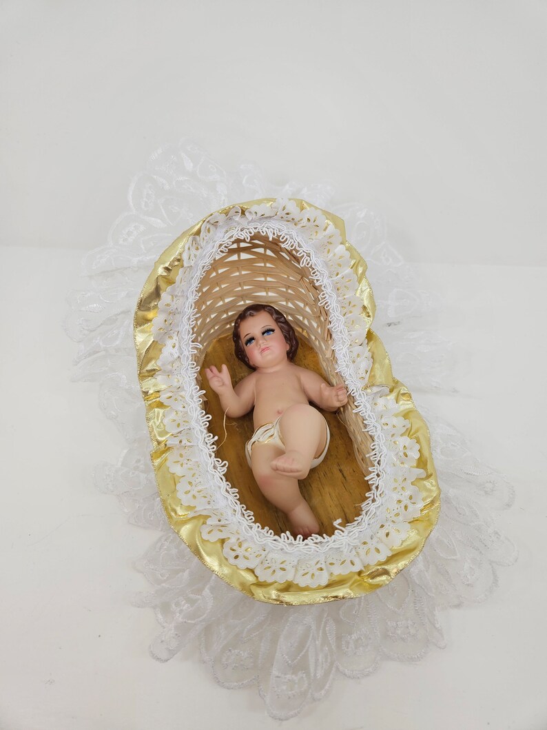K&ouml;nnte beinhalten: Eine kleine Babyfigur mit braunen Haaren und blauen Augen, die in einer geflochtenen Weidenwiege liegt. Die Wiege ist mit gl&auml;nzendem goldfarbenem Stoff ausgekleidet und mit wei&szlig;er Spitze verziert. Das Baby tr&auml;gt eine wei&szlig;e Windel auf einem Holzboden.