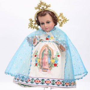 Puede incluir: Una mu&ntilde;eca de porcelana blanca vestida con un vestido blanco con una capa azul y una corona dorada. El vestido tiene una imagen bordada de colores de la Virgen Mar&iacute;a en la parte delantera.