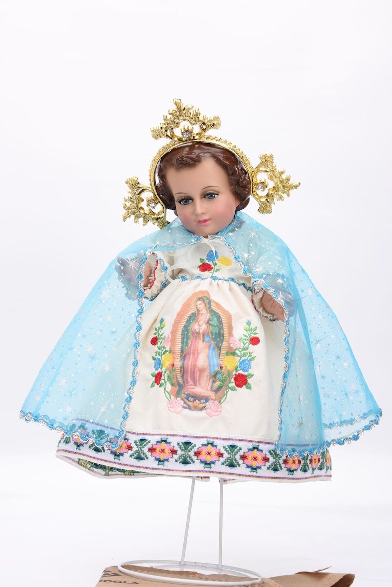 Puede incluir: Una mu&ntilde;eca de porcelana blanca vestida con un vestido blanco con una capa azul y una corona dorada. El vestido tiene una imagen bordada de colores de la Virgen Mar&iacute;a en la parte delantera.