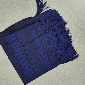 May include: A folded, dark blue scarf with intricate geometric and floral patterns woven throughout. The scarf features a fringe of dark blue tassels at one end. The design incorporates shades of blue and black.