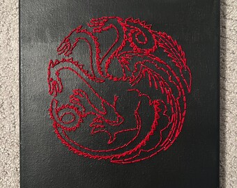 Puede incluir: Un lienzo negro con una imagen bordada en rojo de tres dragones entrelazados. Los dragones están en forma circular y son un símbolo popular de la serie de televisión Game of Thrones.