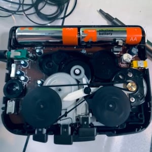 May include: An open, black portable cassette player with its internal components exposed. Two AA alkaline batteries are visible, along with gears, belts, and a circuit board. A small screwdriver rests nearby.