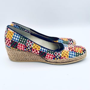 May include: A pair of wedge heel espadrille shoes. The shoes feature a patchwork design with red, yellow, blue, and white floral and gingham patterns. The wedge heels are made of woven jute material, and the shoes have a navy blue trim.