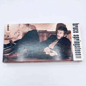 May include: A Bruce Springsteen "Tracks" CD box set. The album cover features a sepia-toned image of a man in a suit reclining on a couch. The artist's name and album title are printed in white on the right side.