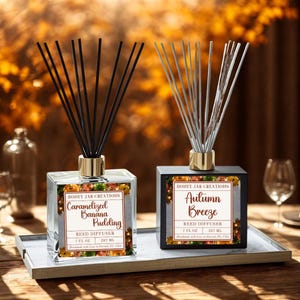 May include: Two reed diffusers on a tray. One is clear with a gold top and black reeds, labeled "Caramelized Banana Pudding." The other is black with silver reeds, labeled "Autumn Breeze." Both have decorative labels.