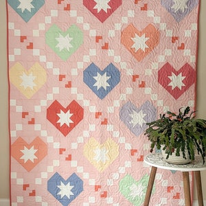 Pode incluir: Uma colcha rosa com um design de patchwork que apresenta corações em várias cores, incluindo vermelho, laranja, amarelo, verde, azul e roxo. Cada coração tem uma estrela branca no centro. A colcha está pendurada em uma parede com uma mesa pequena e uma planta na frente.