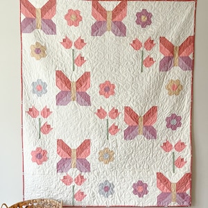 May include: A white quilt featuring a pink and white butterfly and flower design. The quilt is crafted from a range of fabrics and textures.