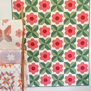 Könnte beinhalten: Eine große Steppdecke mit floralem Muster, die rosa Blütenformen und grüne Sternmuster auf weißem Hintergrund aufweist. Die Steppdecke wird an einer weißen Wand ausgestellt. Andere Steppdecken mit verschiedenen Mustern sind auf einer Holzleiter zu sehen.