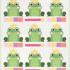 Puede incluir: Un patrón de colcha con nueve ranas verdes que llevan coronas doradas. Cada rana está rodeada de una fila de flores rosas, amarillas, moradas y naranjas. El fondo es blanco.