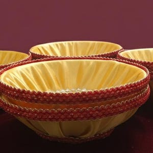 May include: Four decorative bowls with a yellow satin interior and a red beaded trim. The bowls are set on a burgundy surface, with a matching burgundy background.