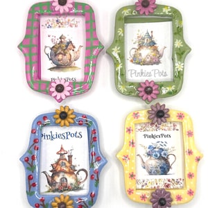 May include: Four rectangular picture frames with rounded, scalloped edges, each decorated with a flower at the top and bottom. The frames are painted in pink plaid, green with white flowers, blue with red berries, and yellow with pink flowers, all displaying PinkiesPots art.
