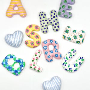May include: A collection of ceramic-style letters and heart shapes featuring various hand-painted patterns. The letters include A, S, H, E, U, L, and B, decorated with stripes, polka dots, floral motifs, and plaid designs in yellow, pink, blue, and green.