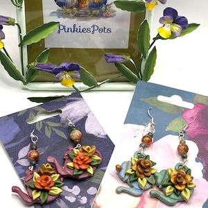May include: Two pairs of handmade polymer clay earrings featuring teapot designs with floral accents. One pair is in shades of green and yellow, while the other pair is in shades of purple and orange.