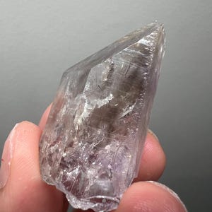 May include: A clear, faceted crystal with a purple hue. The crystal is shaped like a point and has a rough, natural texture.