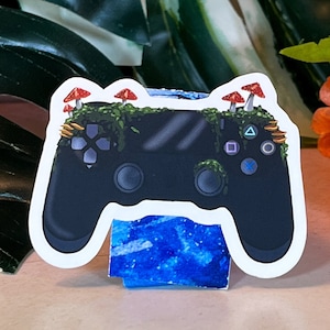 Puede incluir: Una pegatina negra de mando de videojuegos con un diseño de bosque en la parte superior, con setas rojas y musgo verde. El mando está apoyado en un soporte azul y blanco, con un fondo de hojas verdes y flores naranjas.