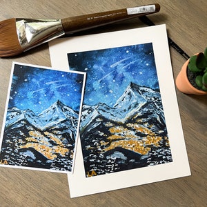 May include: A watercolor painting of a snowy mountain range under a starry night sky. The mountains are painted in shades of blue, white, and black, with hints of gold in the foreground. The sky is a deep blue with white stars and a faint white streak.