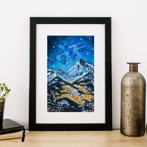 May include: A framed watercolor painting of a mountain range under a starry night sky. The mountains are painted in shades of blue and white, with a hint of gold in the foreground. The sky is a deep blue with white stars and a swirling galaxy.