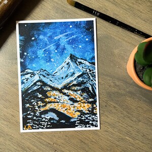 May include: A watercolor painting of a snowy mountain range under a starry night sky. The mountains are painted in shades of blue and white, with a touch of yellow at the base. The sky is a deep blue with white stars and a few streaks of white clouds.