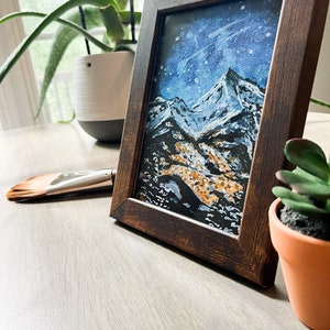 May include: A framed painting of a snowy mountain range with a blue and white sky. The painting is in a dark brown frame and is sitting on a wooden table.