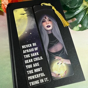May include: A black and white bookmark with a witch holding a glowing orb. The text on the bookmark reads "Never be afraid of the dark dear child, you are the most powerful thing in it."