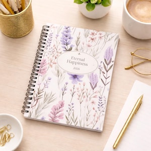 May include: A floral-patterned spiral-bound planner with a cover that reads "Eternal Happiness 2026". The design features purple, pink, and green botanical illustrations. A gold pen and other stationery items are visible on the desk.
