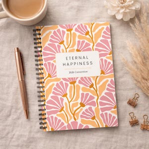 May include: A spiral-bound notebook with a floral pattern in pink and orange. The cover features the text "ETERNAL HAPPINESS 2026 Convention". A rose gold pen and a cup of coffee are next to the notebook. Gold binder clips are also visible.