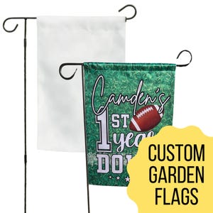 May include: Two garden flags on black metal stands. One flag is white, the other is green with a football design and the text "Camden's 1st year". A yellow sticker reads "Custom Garden Flags".