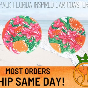 May include: Two round car coasters with a floral and citrus design. The coasters feature pink flowers, green leaves, and orange citrus fruit. The coasters are on a white sandy beach with blue water in the background. The text "2 PACK FLORIDA INSPIRED CAR COASTERS" is at the top of the image. The text "MOST ORDERS SHIP SAME DAY!" is at the bottom of the image.