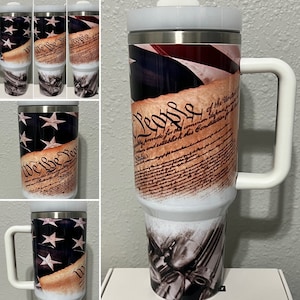 May include: A stainless steel travel mug with a white lid and handle. The mug features a design of the American flag and the text "We the People" from the US Constitution.