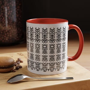 May include: White ceramic mug with a red interior and handle. The mug features a repeating black abstract design. A silver spoon and a macaron are on a wooden surface next to the mug.