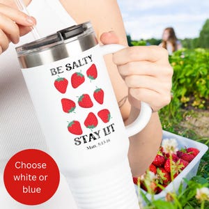 以下が含まれることがあります： 黒い蓋とハンドルが付いた白いトラベルマグ。テキスト「BE SALTY STAY LIT」と赤いイチゴのイラストが特徴です。マグには「Matt. 5:13-16」のテキストもあります。テキスト「Choose white or blue」の赤い円が左下にあります。