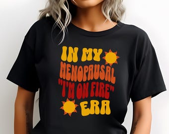 May include: Black t-shirt with the text "IN MY MENOPAUSAL 'I'M ON FIRE' ERA" in orange and yellow. The design includes sun graphics. The shirt is a casual wear item.