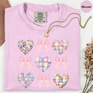 Peut inclure: Un t-shirt rose clair avec une étiquette Comfort Colors, orné de cœurs en patchwork et de nœuds roses. Un collier pendentif étoile doré est drapé sur le t-shirt. Le t-shirt est de taille XL-TG-EG.