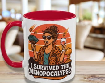 May include: White ceramic mug with a red interior and handle. The mug features a humorous illustration of a woman holding a fan and a wine glass, with the text "I Survived the Menopocalypse."