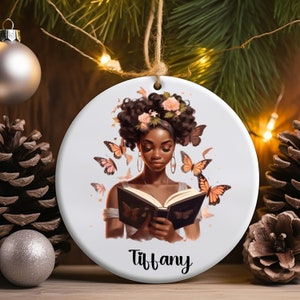 May include: A white ceramic ornament with a colorful illustration of a woman with an afro reading a book. Butterflies surround her and she is wearing a flower crown. The ornament is personalized with the name "Tiffany".