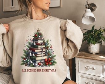 May include: Beige sweatshirt featuring a Christmas tree design made of books, adorned with ornaments, bells, and a star. The text "ALL BOOKED FOR CHRISTMAS" is printed at the bottom. A festive holiday-themed apparel item.