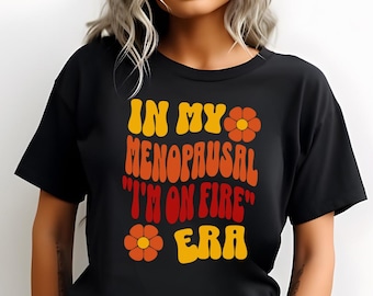 May include: Black t-shirt with the text "IN MY MENOPAUSAL 'I'M ON FIRE' ERA" in yellow, orange, and red. The design includes flower graphics. The shirt has short sleeves and a crew neck.