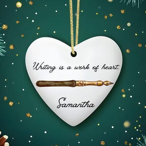 May include: White heart-shaped ornament with the text "Writing is a work of heart" and the name "Samantha". A detailed illustration of a vintage pen is below the text. The ornament hangs from a gold string, set against a festive green background.