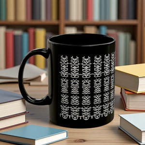 May include: A black ceramic mug with a white decorative pattern and the text "More Chapter". The mug has a large handle and is surrounded by books on a wooden surface.