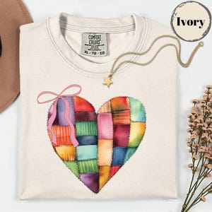 Peut inclure: T-shirt ivoire avec un motif de cœur patchwork coloré et un nœud rose. Un collier en or avec un pendentif en forme d'étoile est posé sur le t-shirt. Le t-shirt porte une étiquette Comfort Colors, taille XL-TG-EG.
