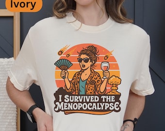 May include: Ivory-colored t-shirt with a graphic featuring a woman in sunglasses holding a fan and a wine glass, with the text "I Survived the Menopocalypse". The design includes a sunset, meteors, and a mushroom cloud.