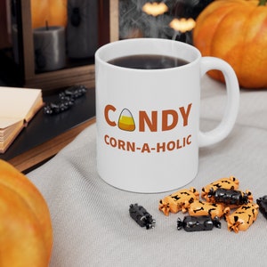 May include: A white ceramic mug with the words "CANDY Corn-a-holic" in orange and a candy corn graphic. The mug is filled with a dark liquid. Halloween-themed sweets are scattered around the mug.