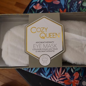以下が含まれることがあります： 箱に入っている白いふわふわのアイマスク。箱には「Cozy Queen」と「Aromatherapy Eye Mask」の文字が書かれている。