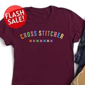 May include: A maroon t-shirt with the text "CROSS STITCHER" in rainbow colors. The text is in a curved font and is followed by a row of colorful "X" symbols.