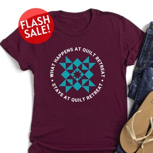 May include: A maroon t-shirt with a white circle design that says "What happens at quilt retreat stays at quilt retreat". The design features a teal quilt block pattern.
